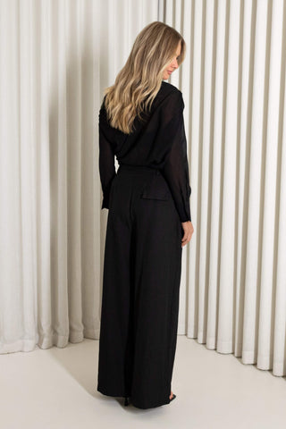 A woman wearing a black long-sleeve jumpsuit with wide legs, shown from the back against a background of white vertical pleated curtains.
