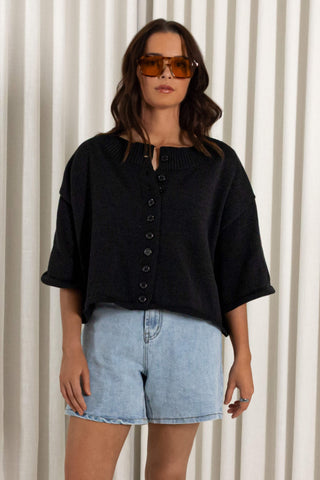 A woman modeling a black short-sleeve button-front knit top with light blue denim shorts, standing in front of a cream vertical-striped curtain backdrop.