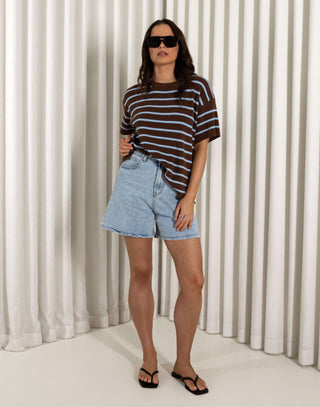Woman wearing a striped shirt and denim shorts standing against a white curtain. Model wearing blue high waisted stretch denim shorts, relaxed fit