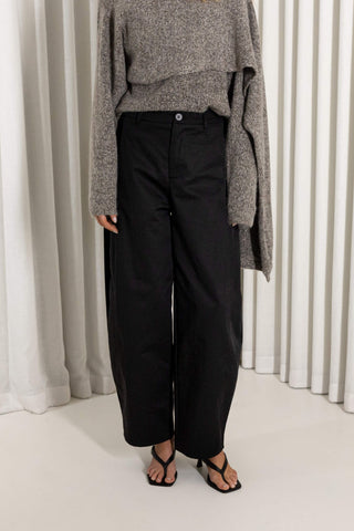 A model wearing black wide-leg cropped trousers, a gray knit sweater, and black strappy sandals, standing in front of a white curtain backdrop.