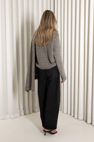 A woman is shown from behind wearing a gray knit sweater, black wide-leg trousers, and black heeled sandals, standing in front of white vertical drapes.