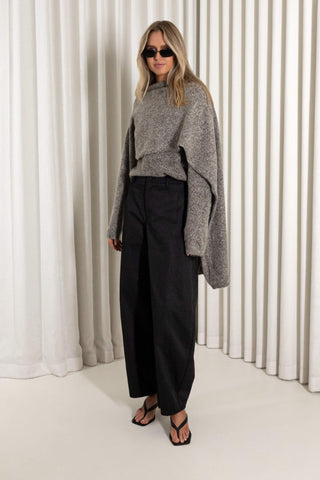 A woman modeling a gray oversized knit sweater and black wide-leg trousers, standing in front of white vertical drapes.