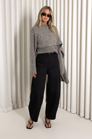 A woman modeling a gray knit draped sweater and black wide-leg trousers, standing in front of a white pleated curtain backdrop.