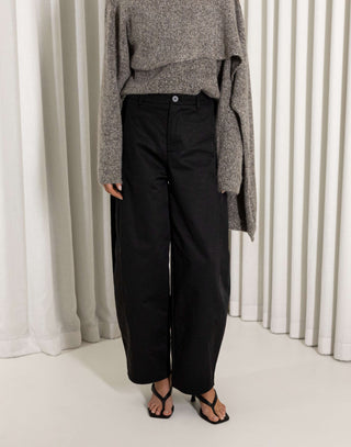 Person wearing a gray sweater and black pants standing against a white curtain background
