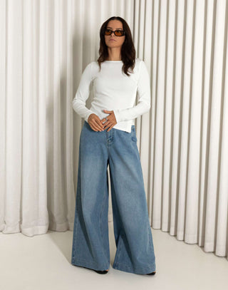 Person wearing a white top and blue jeans standing against a white curtain background high waisted wide leg jeans in blue