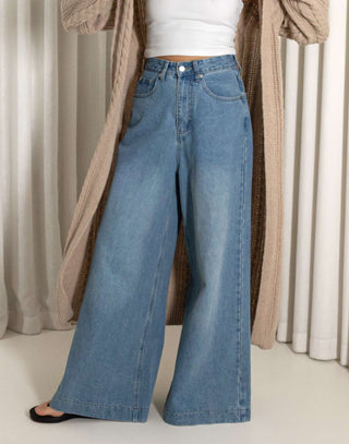 Woman wearing a nude cardigan, white top, and blue jeans against a neutral background high waisted wide leg jeans in blue