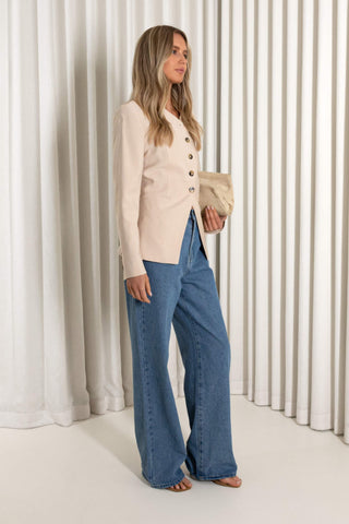 A woman stands in front of white vertical drapes wearing a light beige button-front cardigan, wide-leg blue jeans, and flat sandals, while holding a small beige clutch.