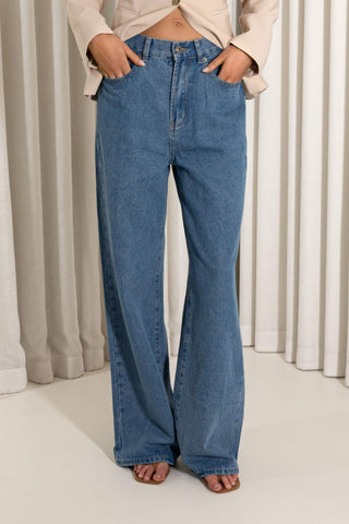 A person wearing loose, wide-leg blue denim jeans with a beige top, standing barefoot in front of beige curtains.