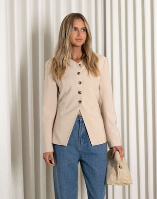 Woman wearing a beige blazer and blue jeans against a white curtain background