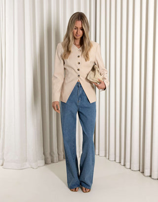 Woman wearing a beige blazer and blue jeans standing against a white curtain.