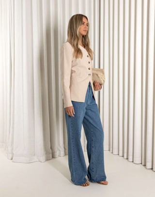 Woman wearing a beige blazer and blue jeans standing against a white curtain.