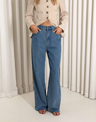 Person wearing blue jeans and a beige jacket standing against a white curtain background