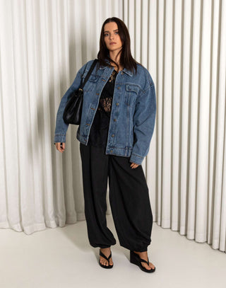 Woman wearing a denim jacket and black pants standing against a white curtain background