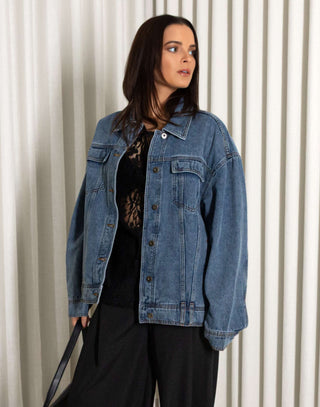 Woman wearing a denim jacket and black pants standing against a white curtain background