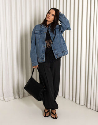 Woman wearing a denim jacket and black pants standing against a white curtain background