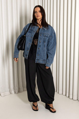 A woman stands in front of a white pleated curtain backdrop wearing an oversized blue denim jacket over a black top and black wide-leg pants, with black sandals and a black shoulder bag.