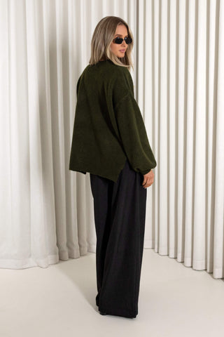A woman wearing a loose olive green cropped sweater and black wide-leg pants, standing in front of white vertical drapes.