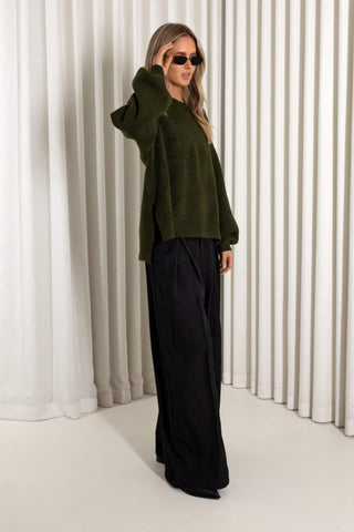 A woman modeling an olive green fuzzy cropped sweater and black wide-leg trousers, standing in front of white vertical drapes.