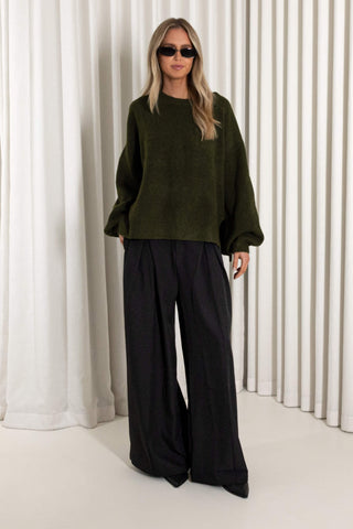 A woman modeling an oversized dark olive green knit sweater with voluminous long sleeves, paired with black wide-leg trousers, standing in front of a white pleated curtain backdrop.
