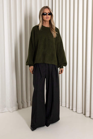 A woman modeling an oversized dark olive green knit sweater and black wide-leg trousers, standing in front of a white curtain backdrop.