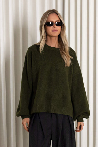 A woman modeling an oversized olive green knit sweater with voluminous long sleeves, paired with dark trousers and black sunglasses, standing against a white curtain backdrop.