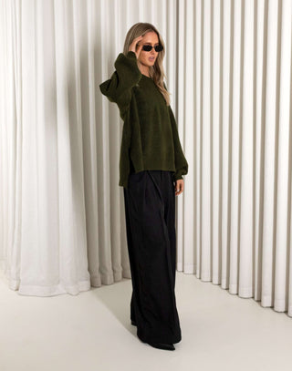 Person wearing an olive green sweater and black pants against a white curtain background