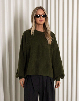 Woman wearing a green sweater and black pants against a white curtain background
