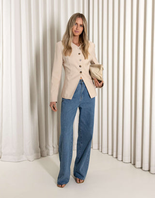 Woman wearing a beige cardigan and blue jeans standing against a white curtain background