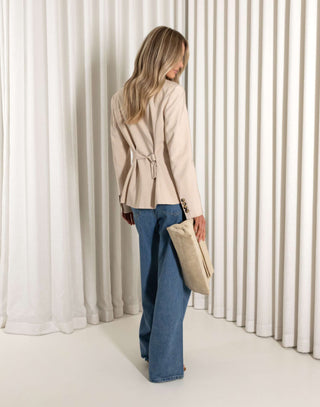 Woman wearing a beige blazer and blue jeans holding a beige bag against a white curtain background