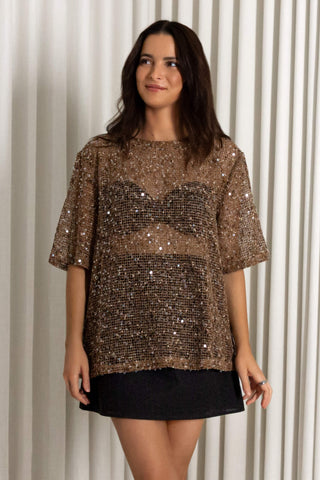 A woman modeling a short-sleeved sequined top with a sheer mesh chest panel and a black mini skirt, standing in front of a cream-colored pleated curtain backdrop.