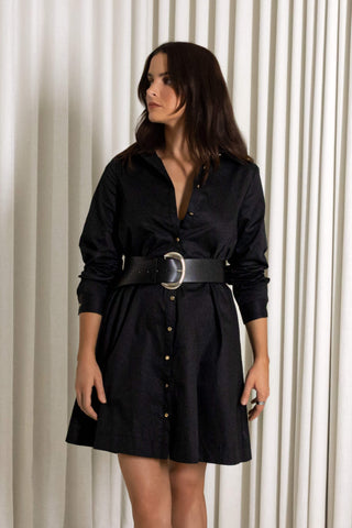 A woman wearing a black long-sleeve button-front shirt dress with a wide black belt and silver buckle, standing in front of a cream-colored pleated curtain backdrop.