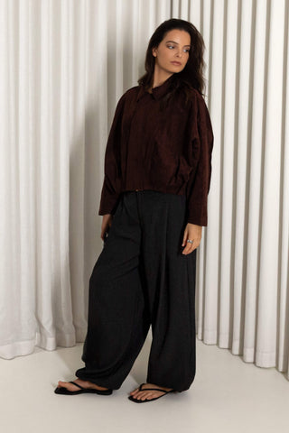 A woman modeling a dark brown long-sleeve collared shirt with black wide-leg trousers and black sandals, standing in front of white vertical drapes.
