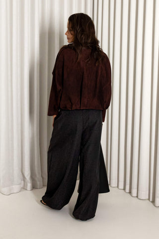 A woman stands with her back to the camera wearing a dark brown cropped jacket and wide-leg black trousers, posed in front of white vertical drapes.
