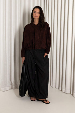 A woman stands in front of white pleated curtains wearing a dark brown long-sleeve collared shirt, black wide-leg pants, and black sandals, holding a black bag.