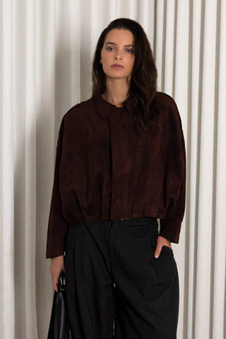 A woman modeling a dark burgundy long-sleeve textured blouse with a collar, paired with black wide-leg trousers, standing in front of a white pleated curtain backdrop.