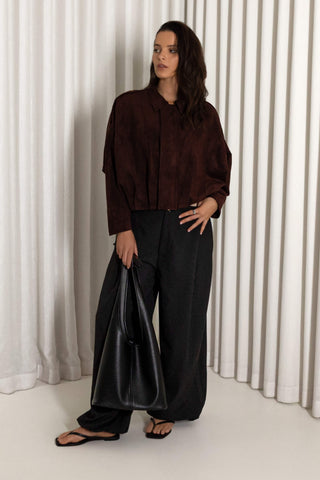 A woman stands in a studio wearing a dark brown long-sleeve collared shirt, loose black wide-leg pants, and black sandals, while holding a large black tote bag. She is posed in front of white vertical drapes.