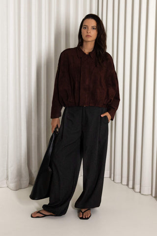 A woman stands in front of white vertical drapes wearing a dark brown long-sleeve button-up shirt, black wide-leg trousers, and black sandals, holding a black bag.