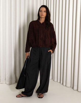 Person wearing a dark brown faux suede bomber and black balloon pants standing against a white curtain.