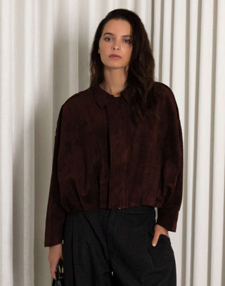 Person wearing a dark brown faux suede bomber and black balloon pants standing against a white curtain.