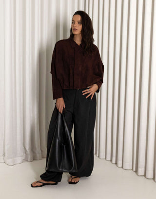 Person wearing a dark brown faux suede bomber and black balloon pants standing against a white curtain.
