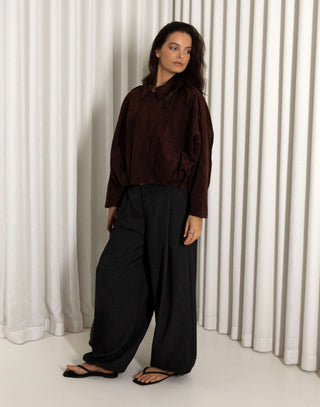 Person wearing a dark brown faux suede bomber and black balloon pants standing against a white curtain.