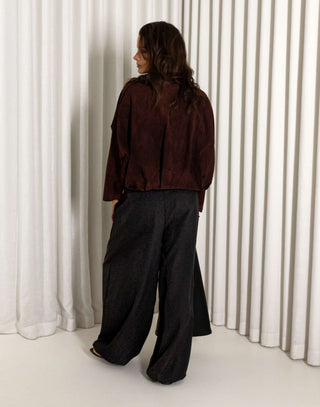 Person wearing a dark brown faux suede bomber and black balloon pants standing against a white curtain.