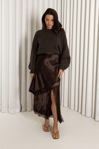 A woman stands in a studio wearing a dark brown knit sweater and a matching dark brown satin midi skirt with a lace hem, holding a small dark handbag.