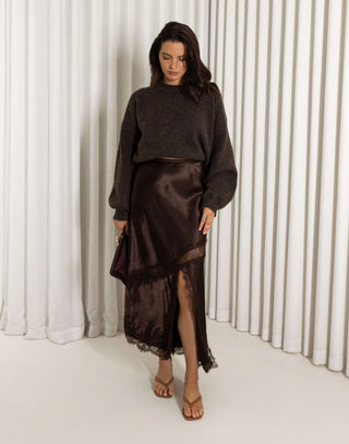 Woman wearing a dark brown sweater and brown lace midi skirt with front split against a white curtain background
