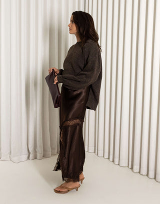 Woman wearing a dark brown sweater and brown lace midi skirt with front split against a white curtain background