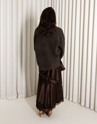 Woman wearing a dark brown sweater and brown lace midi skirt with front split against a white curtain background