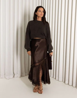 Woman wearing a dark brown sweater and brown lace midi skirt with front split against a white curtain background