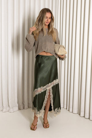 A woman stands in front of white pleated curtains wearing a taupe cropped knit sweater and a dark olive green satin midi skirt with a lace hem. She is holding a beige clutch and wearing tan flat sandals.