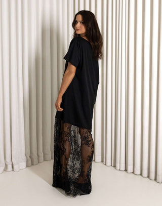 Woman wearing a black one-shoulder dress with lace detailing against a white curtain background Black off shoulder lace maxi dress
