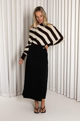 A woman modeling a brown and cream striped off-the-shoulder knit top paired with a long black skirt and black thong sandals, standing in front of a white pleated curtain backdrop.
